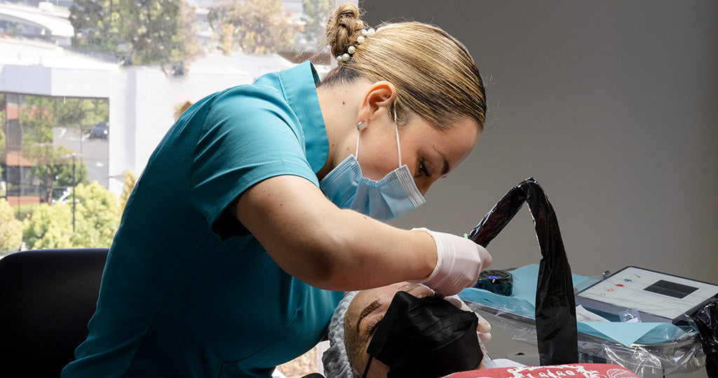 Cosmetic professional performing a cosmetic eyebrow procedure on a client in a treatment setting
