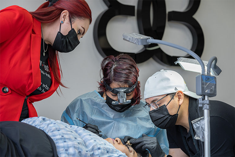 Three people in a medical or tattoo setting with a butterfly logo in the background.